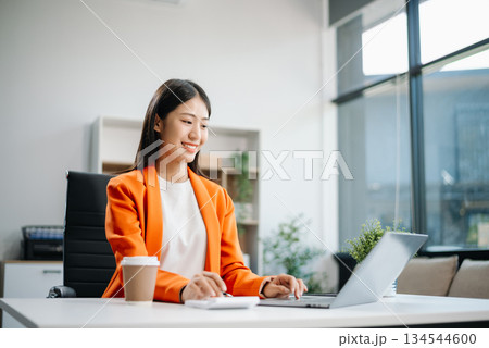 Asian woman, smiling and working at her desk in an office environment. She is wearing a business suit and holding documents while using a laptop. Asian woman, smiling and working at her desk in an office environment. She is wearing a business suit and holding documents while using a laptop. 134544600