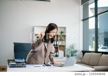 Asian woman, smiling and working at her desk in an office environment. She is wearing a business suit and holding documents while using a laptop. 134544646