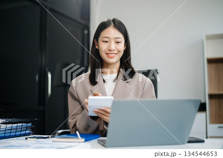 Asian woman, smiling and working at her desk in an office environment. She is wearing a business suit and holding documents while using a laptop. 134544653
