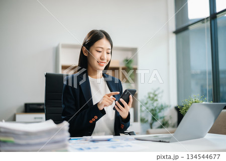 Young Asian woman, smiling and working at her desk in an office environment. She is wearing a business suit and holding documents Young Asian woman, smiling and working at her desk in an office environment. She is wearing a business suit and holding documents 134544677