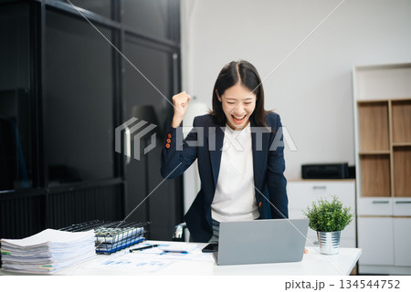 Asian woman, smiling and working at her desk in an office environment. She is wearing a business suit and holding documents while using a laptop. 134544752