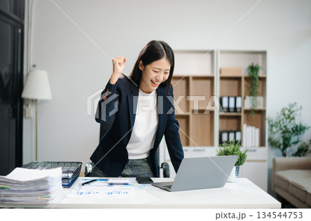 Asian woman, smiling and working at her desk in an office environment. She is wearing a business suit and holding documents while using a laptop. Asian woman, smiling and working at her desk in an office environment. She is wearing a business suit and holding documents while using a laptop. 134544753