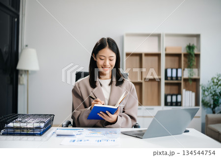 Asian woman, smiling and working at her desk in an office environment. She is wearing a business suit and holding documents while using a laptop. Asian woman, smiling and working at her desk in an office environment. She is wearing a business suit and holding documents while using a laptop. 134544754