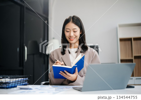 Asian woman, smiling and working at her desk in an office environment. She is wearing a business suit and holding documents while using a laptop. 134544755