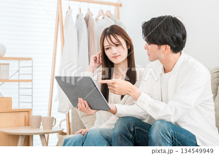 家のリビングでソファーに座りタブレットを見る若いアジア人夫婦,カップル 134549949