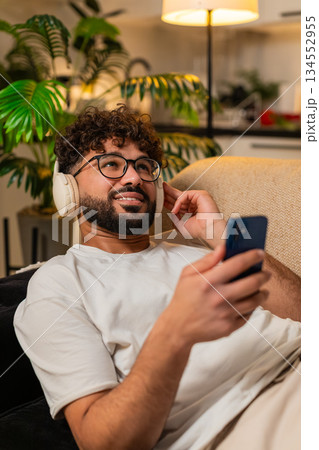 Arabian man wearing wireless headphones listens music texts on smartphone at night indoors relaxed Arabian man wearing wireless headphones listens music texts on smartphone at night indoors relaxed 134552955