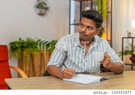 Indian man writes goals in notebook sips coffee ticks boxes smiles and plans day focused at desk 134552971