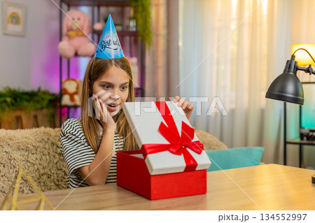 Child girl kid celebrates birthday happily opens gift with excited surprise at table on home sofa Child girl kid celebrates birthday happily opens gift with excited surprise at table on home sofa 134552997