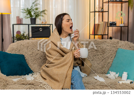 Young woman wrapped in plaid blanket sitting on sofa holding hot tea feeling sick with cold symptoms 134553002
