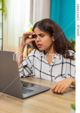 Indian woman holding head confused at laptop screen trying struggling with big complex issue indoors Indian woman holding head confused at laptop screen trying struggling with big complex issue indoors 134553082