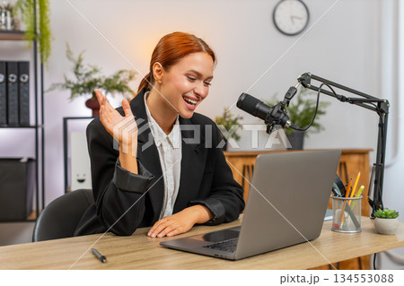 Young businesswoman records podcast blog speaking to studio microphone beside laptop laughing loudly 134553088
