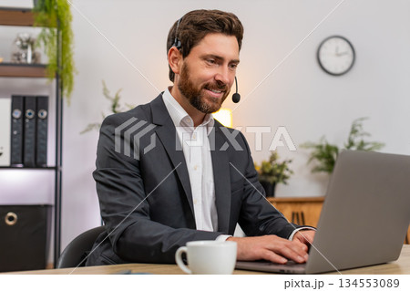 Middle-aged businessman service agent on headset listens and solves client issue while typing notes Middle-aged businessman service agent on headset listens and solves client issue while typing notes 134553089