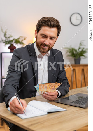 Middle-aged businessman counts cash euros enters budget on laptop writes notes in notebook and plans Middle-aged businessman counts cash euros enters budget on laptop writes notes in notebook and plans 134553128