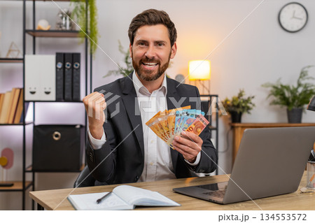 Middle-aged businessman counts cash euros enters budget on laptop writes notes in notebook and plans Middle-aged businessman counts cash euros enters budget on laptop writes notes in notebook and plans 134553572