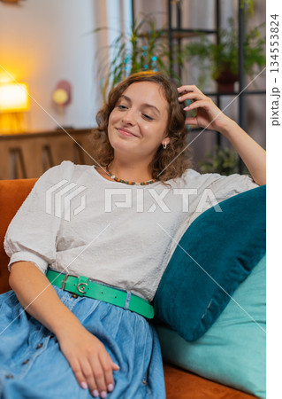 Happy carefree young Caucasian woman sitting comfortably on sofa thinking in living room at home Happy carefree young Caucasian woman sitting comfortably on sofa thinking in living room at home 134553824