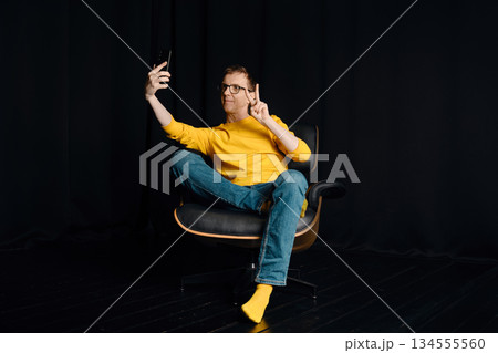 Middle-aged man sits on chair and takes selfie in dark room with yellow shirt and blue jeans 134555560