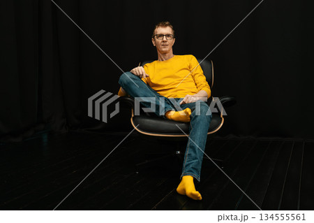 Middle-aged man sits comfortably in a chair wearing a yellow sweater and socks in a dark room Middle-aged man sits comfortably in a chair wearing a yellow sweater and socks in a dark room 134555561