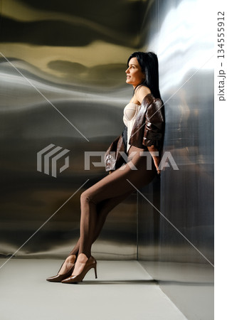 Woman poses in studio against shiny background wearing fitted outfit and high heels Woman poses in studio against shiny background wearing fitted outfit and high heels 134555912