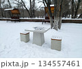 A concrete outdoor chessboard table with two matching stools, dusted with snow, sits in a snowy park setting with bare trees and blurred structures in the background 134557461