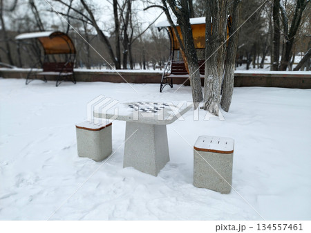 A concrete outdoor chessboard table with two matching stools, dusted with snow, sits in a snowy park setting with bare trees and blurred structures in the background A concrete outdoor chessboard table with two matching stools, dusted with snow, sits in a snowy park setting with bare trees and blurred structures in the background 134557461
