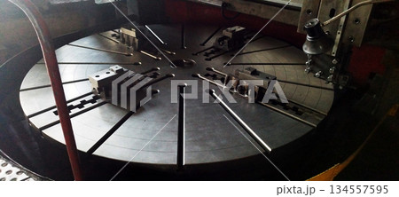The massive, slotted rotating table of a heavy-duty vertical turning lathe, featuring several large metal chuck jaws, sits under dim industrial lighting The massive, slotted rotating table of a heavy-duty vertical turning lathe, featuring several large metal chuck jaws, sits under dim industrial lighting 134557595