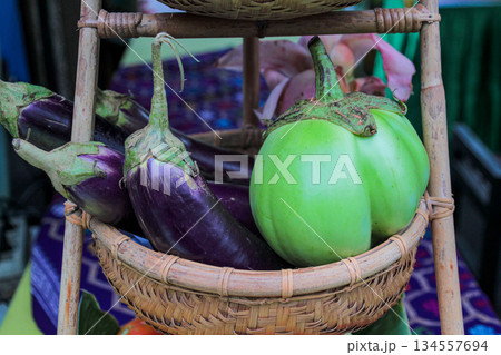 Macro shot capturing the texture and contrast of raw, organically grown Solanum melongena (purple eggplant) next to a round green Thai eggplant variety (Solanum xanthocarpum) in a rattan carrier. 134557694