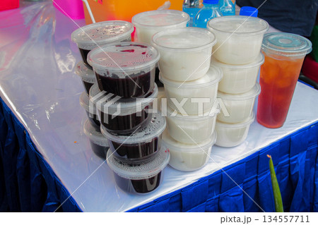 High-angle medium shot showing plastic cups stacked with Bubur Ketan Hitam (Black Glutinous Rice Porridge) and white pudding/sumsum, displayed for sale at a traditional food market event. 134557711