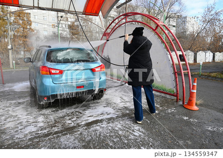 Woman washes her car using a pressure washer at a self-service car wash in the city during a gray day Woman washes her car using a pressure washer at a self-service car wash in the city during a gray day 134559347