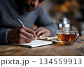 man writing in a wellness journal at a wooden table, cup of herbal tea nearby 134559913