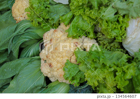 Close-up view of fresh cauliflower head surrounded by assorted green lettuce and leafy vegetables at market stall. 134564607
