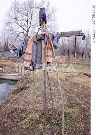 There stands a scarecrow. It is made of a wooden cross, over which is draped a blue cap and an orange vest, imitating a person. The scarecrow stands in a field, next to a pond with calm water. There stands a scarecrow. It is made of a wooden cross, over which is draped a blue cap and an orange vest, imitating a person. The scarecrow stands in a field, next to a pond with calm water. 134565310