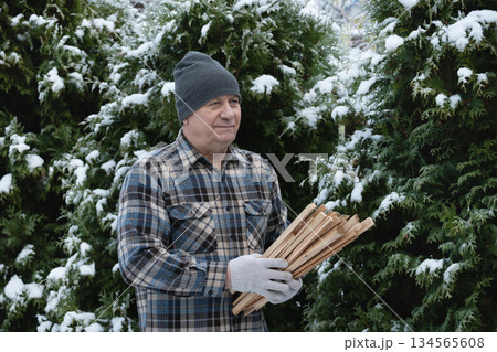Senior man carrying firewood outside in snowy backyard. Practical winter preparation for home heating. Realistic lifestyle image for energy, fuel, and rural living concepts. Senior man carrying firewood outside in snowy backyard. Practical winter preparation for home heating. Realistic lifestyle image for energy, fuel, and rural living concepts. 134565608