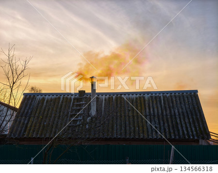 House roof with a metal chimney releasing colorful smoke against a vibrant orange and yellow sunset sky, symbolizing warmth, home comfort, and potential air pollution in a rural winter setting. House roof with a metal chimney releasing colorful smoke against a vibrant orange and yellow sunset sky, symbolizing warmth, home comfort, and potential air pollution in a rural winter setting. 134566318