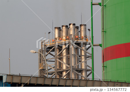 Five industrial metal exhaust stacks supported by a steel framework next to a bright green storage tank structure against a gray sky. Five industrial metal exhaust stacks supported by a steel framework next to a bright green storage tank structure against a gray sky. 134567772