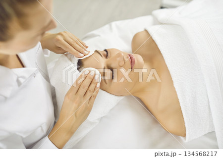 Beautician uses cotton pads with cleanser to remove makeup from face of happy young woman 134568217