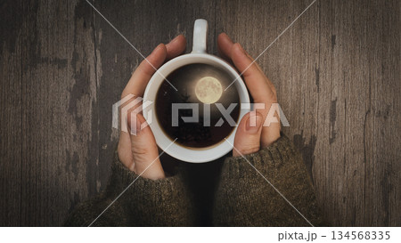 Female hands in a green sweaters sleeves with coffee cup on wooden table, top view. A full Moon and black night forest in the cup. Female hands in a green sweaters sleeves with coffee cup on wooden table, top view. A full Moon and black night forest in the cup. 134568335