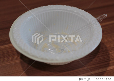 White disposable plastic bowl with residual food and clear plastic spoon resting on brown wooden table after finishing meal. White disposable plastic bowl with residual food and clear plastic spoon resting on brown wooden table after finishing meal. 134568372