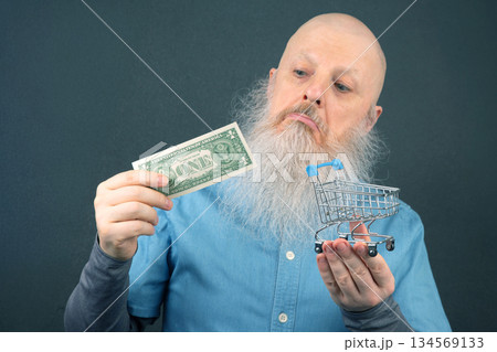 bald man with a large white beard holds a dollar and a model of a grocery basket 134569133