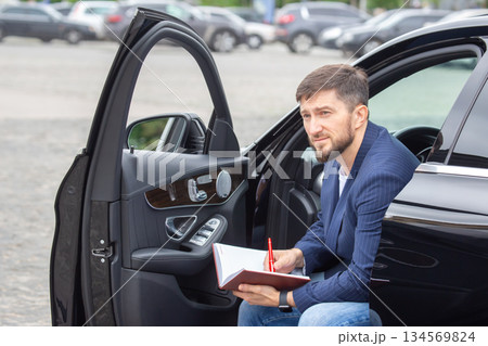Man sits in car with notebook and pen in urban parking lot during daylight Man sits in car with notebook and pen in urban parking lot during daylight 134569824