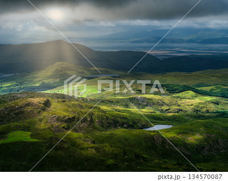 Sunlight shines through clouds onto green hills near the Coomloughra Horseshoe trail. Mountains fade into the distance. Small lakes dot the landscape in County Kerry, Ireland. Sunlight shines through clouds onto green hills near the Coomloughra Horseshoe trail. Mountains fade into the distance. Small lakes dot the landscape in County Kerry, Ireland. 134570087
