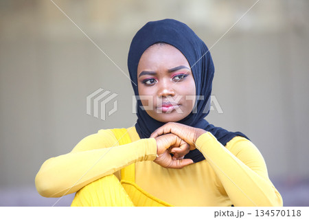 Young african Woman in a Black Hijab and Yellow Dress Reflecting Thoughtfully in an Outdoor Setting 134570118