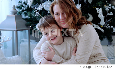 Mother and son embrace in front of the holiday tree, capturing a heartwarming moment of love and unity during the festive season. A day with family before Christmas. New Year's Eve with family Mother and son embrace in front of the holiday tree, capturing a heartwarming moment of love and unity during the festive season. A day with family before Christmas. New Year's Eve with family 134570182