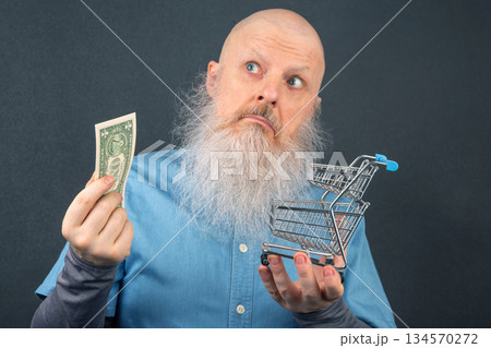 Man with a beard holding dollar bill and shopping cart contemplating spending choices Man with a beard holding dollar bill and shopping cart contemplating spending choices 134570272