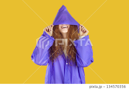 Smiling curly redhead girl in purple raincoat hiding in hood on her eyes on yellow background. 134570356
