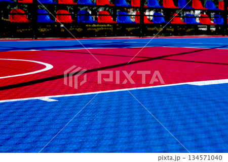Vibrant basketball court with red and blue colors under bright sunlight, showing court markings and shadows during daytime activity 134571040