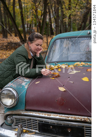 Woman leans on a rusty vintage car, touching yellow autumn leaves on the hood, with a quiet forest and muted fall colors around Woman leans on a rusty vintage car, touching yellow autumn leaves on the hood, with a quiet forest and muted fall colors around 134571844