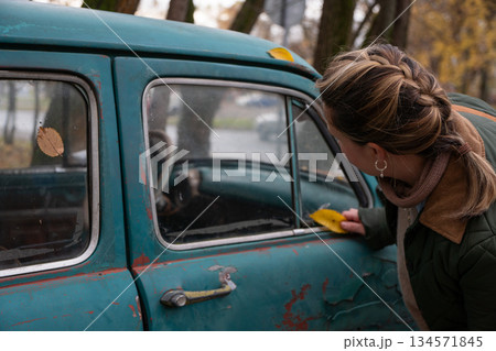 Vintage car with peeling turquoise paint and rusted details stands in quiet autumn courtyard as woman gently touches the window, fallen yellow leaves 134571845