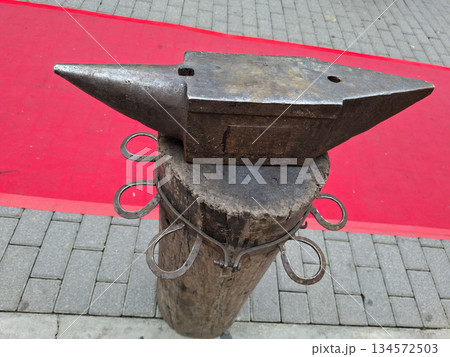 An antique anvil rests on a wooden post, ready for use in hand forging metal. The scene shows a red carpet or mat in the background, indicating a special event or display. 134572503