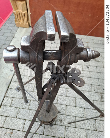 An antique vise stands on a sturdy base, ready for use in metal forging. This tool holds metal securely while craftsmen shape and work on projects. It is a key part of the workshop. 134572504