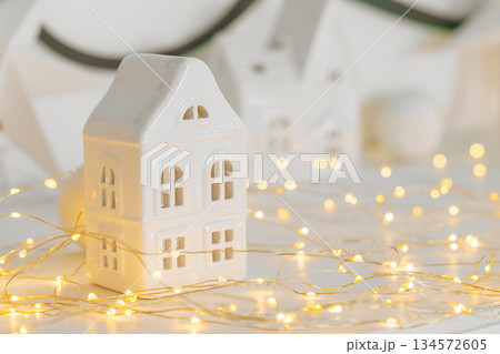 Minimalist Christmas decor, ceramic house on garland warm festive fairy lights background Minimalist Christmas decor, ceramic house on garland warm festive fairy lights background 134572605